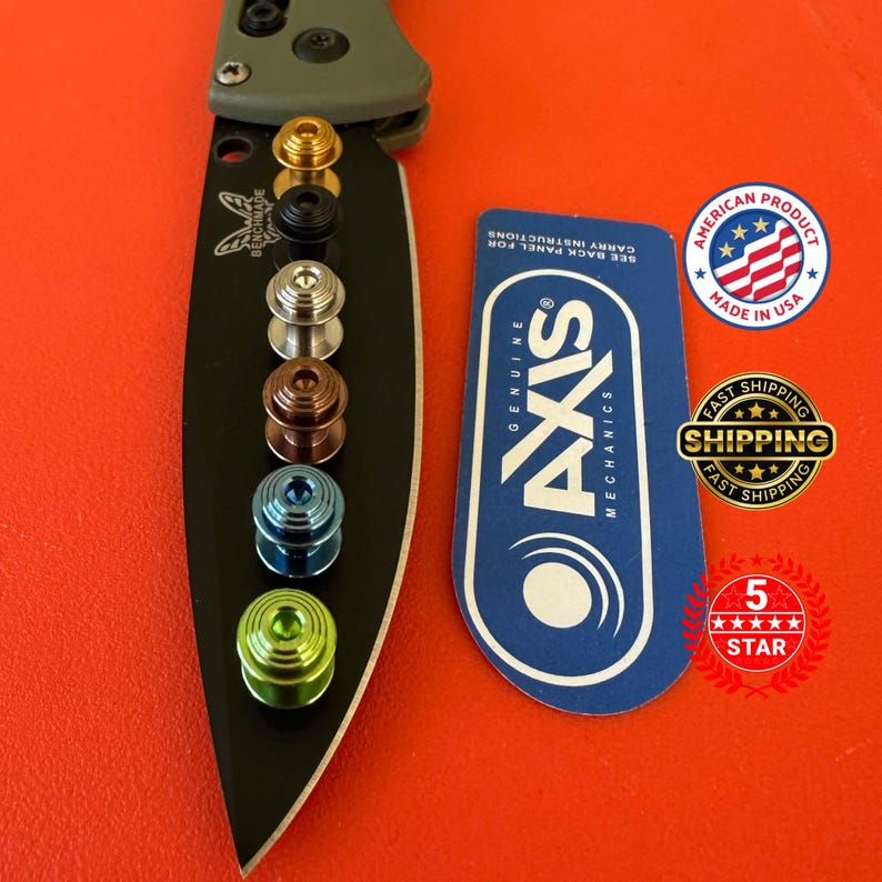 May include: A black Benchmade knife blade with a green handle, showcasing a series of colorful Axis lock thumb studs in gold, black, silver, bronze, blue, and green. A blue tag with "AXIS MECHANICS" text and "MADE IN USA" sticker are also visible.