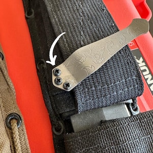 May include: A silver-colored pocket knife clip with an engraved dragon design, attached to a black nylon sheath. The clip is secured with three black screws. The background includes a red container and a tan sheath.