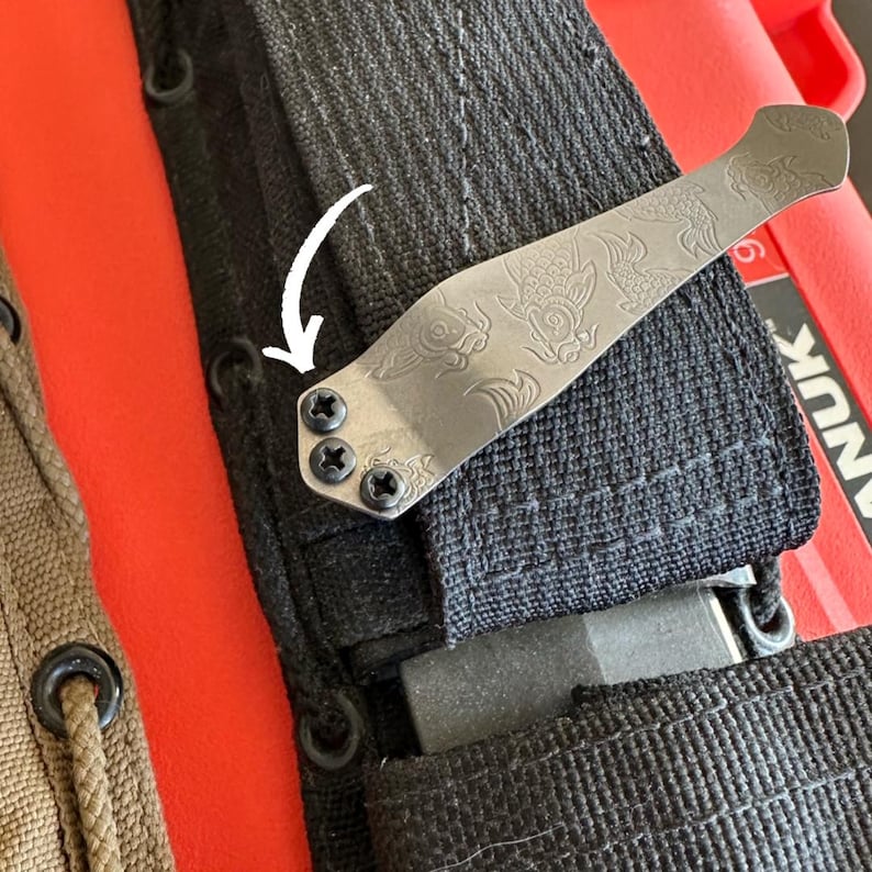 May include: A silver-colored pocket knife clip with an engraved dragon design, attached to a black nylon sheath. The clip is secured with three black screws. The background includes a red container and a tan sheath.