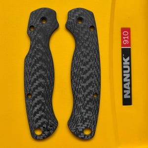 May include: Two black carbon fiber knife handle scales with a textured pattern, featuring three holes each for attachment. A red and black label with the text "NANUK" and "910" is also present.