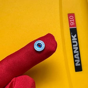以下が含まれることがあります： 赤色の布製のオブジェクトのクローズアップ。小さな円形の青と銀色の金属片が取り付けられています。金属片の縁にはテキストがあります。背景は黄色で、「NANUK」の文字と「910」の数字が印刷されています。