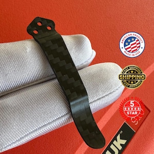 Op de afbeelding: Een zwarte carbon fiber pocketclip met een getextureerd weefpatroon. De clip heeft drie gaten aan de bovenkant en een gebogen onderkant. De afbeelding bevat ook een "Made in USA" sticker.
