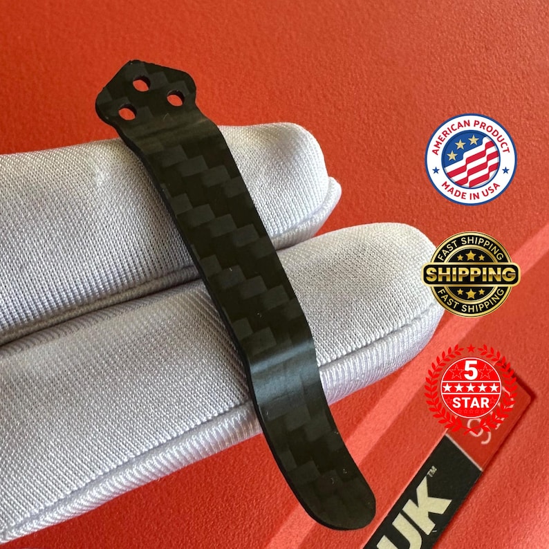 Op de afbeelding: Een zwarte carbon fiber pocketclip met een getextureerd weefpatroon. De clip heeft drie gaten aan de bovenkant en een gebogen onderkant. De afbeelding bevat ook een "Made in USA" sticker.