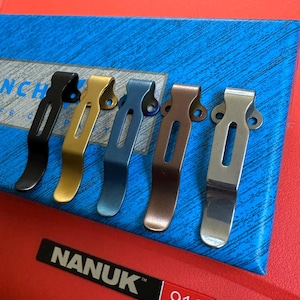 May include: A collection of six metal pocket clips in various colors: black, gold, blue, bronze, and silver. The clips are arranged on a blue surface, with a red label that reads "NANUK".
