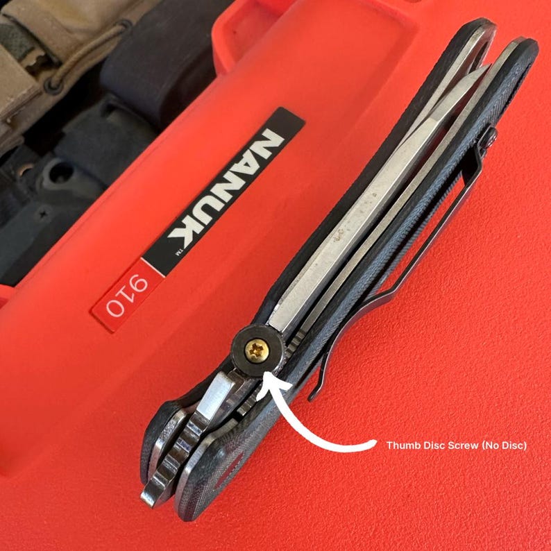May include: A partially open folding knife with a black handle and a silver blade. The knife has a pocket clip and a thumb disc screw. The knife is resting on a red case with the text "NANUK 910".