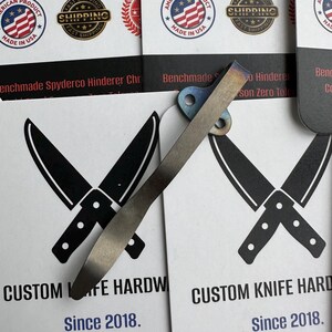 Op de afbeelding: Een close-up van een hardware clip voor een op maat gemaakt mes met een blauw en zilveren afwerking. De clip is geplaatst op een kaart met gekruiste mes illustraties en de tekst "CUSTOM KNIFE HARDWARE Since 2018."