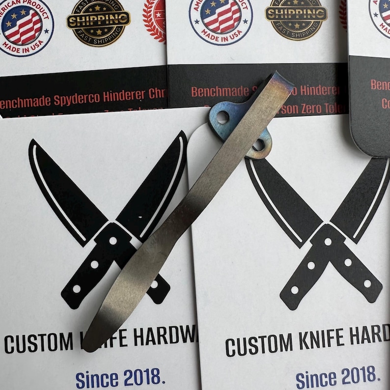 Op de afbeelding: Een close-up van een hardware clip voor een op maat gemaakt mes met een blauw en zilveren afwerking. De clip is geplaatst op een kaart met gekruiste mes illustraties en de tekst "CUSTOM KNIFE HARDWARE Since 2018."