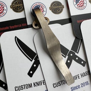 May include: A close-up of a brushed metal pocket clip for a custom knife. The clip is attached to a card with a knife illustration and the text "CUSTOM KNIFE". Other cards feature text like "AMERICAN PRODUCT" and "MADE IN USA".