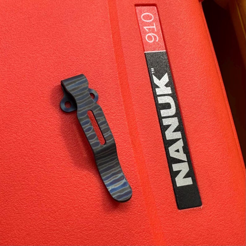 May include: A titanium pocket clip with a blue and black wave pattern, lying on a red surface. The clip has a slotted design and two mounting holes. The word "NANUK" is printed on a black label, and the number "910" is printed on a red label.