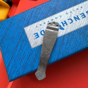 Op de afbeelding: Een zilverkleurige mesclip met een ge&euml;tst visontwerp. De clip is geplaatst op een blauwe doos met de witte tekst "BENCHMADE KNIFE COMPANY". De clip heeft drie gaten aan de bovenkant voor bevestiging.