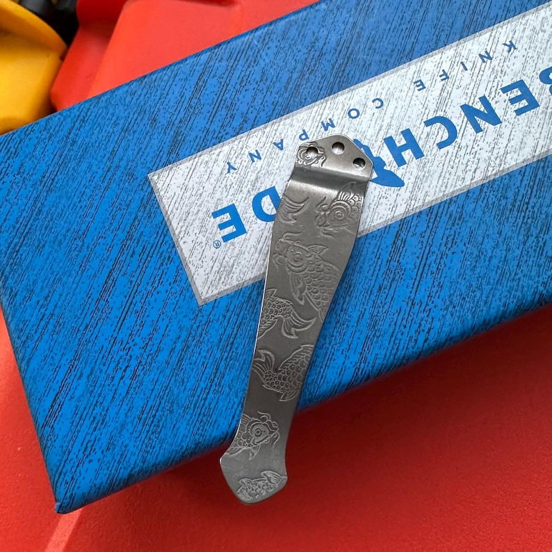 Op de afbeelding: Een zilverkleurige mesclip met een ge&euml;tst visontwerp. De clip is geplaatst op een blauwe doos met de witte tekst "BENCHMADE KNIFE COMPANY". De clip heeft drie gaten aan de bovenkant voor bevestiging.