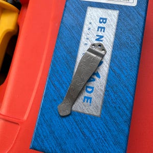 Op de afbeelding: Een zilverkleurige mesclip met een gegraveerd drakenontwerp, rustend op een blauw-witte doos. Op de doos staat de tekst "BENCHMADE KNIFE". De clip is gemaakt van metaal en heeft een gebogen vorm.