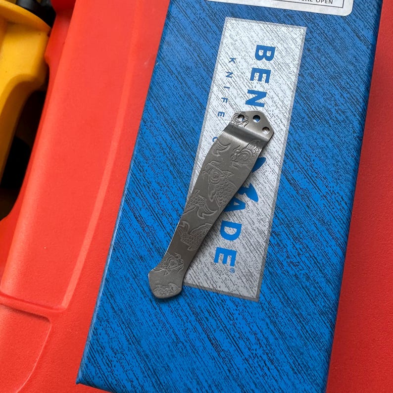 Op de afbeelding: Een zilverkleurige mesclip met een gegraveerd drakenontwerp, rustend op een blauw-witte doos. Op de doos staat de tekst "BENCHMADE KNIFE". De clip is gemaakt van metaal en heeft een gebogen vorm.