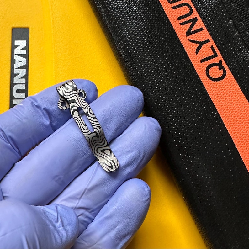 K&ouml;nnte beinhalten: Ein kleiner, gemusterter Metallclip mit einem wellenartigen Design. Der Clip ist silber und schwarz und scheint aus Metall zu sein. Der Hintergrund ist gelb und schwarz.