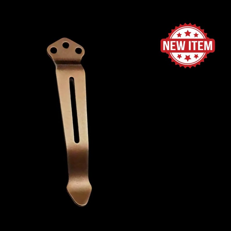 May include: A bronze-colored pocket clip with a slotted design and three small holes at the top. The clip is shown against a black background with a red "NEW ITEM" badge.