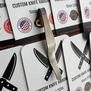May include: A custom knife hardware clip with a brushed metal finish and blue accents. The clip is attached to a small, round base. The background features business cards with knife illustrations and the text "CUSTOM KNIFE HARDWARE Since 2018."
