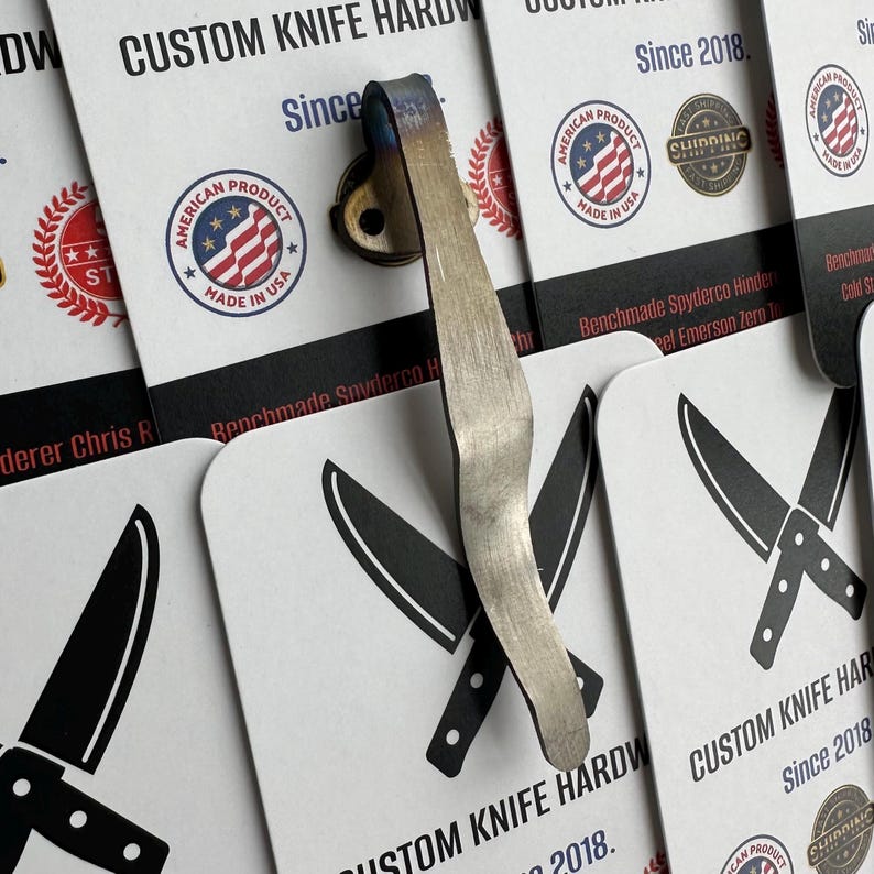 May include: A custom knife hardware clip with a brushed metal finish and blue accents. The clip is attached to a small, round base. The background features business cards with knife illustrations and the text "CUSTOM KNIFE HARDWARE Since 2018."