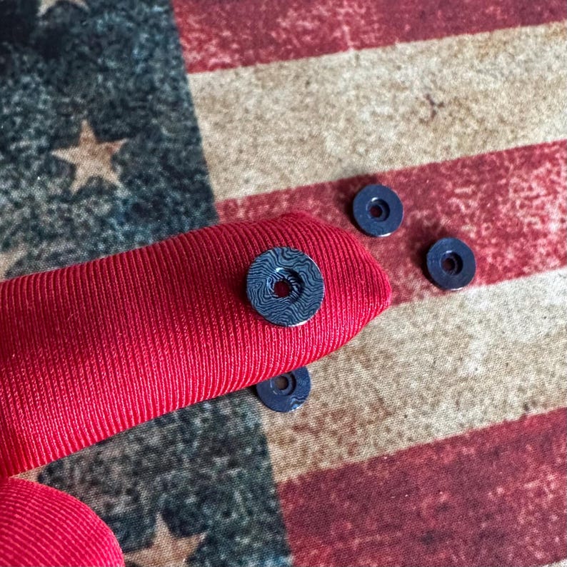 Pu&ograve; includere: Primo piano di diverse piccole rondelle blu scuro con un motivo, appoggiate su un tessuto rosso. Lo sfondo presenta un motivo sbiadito della bandiera americana in rosso, bianco e blu.