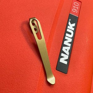 May include: A gold-colored metal clip with two small holes near the top, and a curved bottom. Next to the clip is a black and red label with the word "NANUK" in white, and the number "910". The background is a bright red.