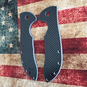 May include: Two carbon fiber knife scales with a checkered pattern. The scales are dark gray with a light gray edge and several holes for screws. The background is an American flag.