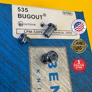 May include: A close-up of several small, metallic blue knife parts, including screws and a cylindrical piece. The parts are arranged on a blue and white patterned surface. Text on the packaging reads "535 BUGUOT®" and "AMERICAN PRODUCT MADE IN USA."
