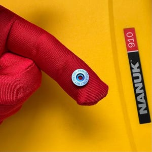 以下が含まれることがあります： 「COLT PISTOL 45 AUTO」と書かれた青と白の文字が入った、小さくて丸い銀色のボタンが赤い布に付いています。背景は黄色で、「NANUK 910」と書かれた黒と赤のラベルが見えます。