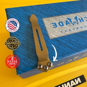 May include: A gold-colored metal pocket clip with three screws, resting on a blue box. The clip has a pointed top and a slotted design. The box has the text "Benchmade Company" and several stickers.