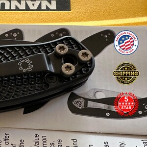 May include: Close-up of a black folding knife with a textured handle and silver screws. The knife features a pocket clip with a logo and is labeled as an American product.