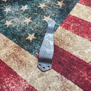 May include: A silver-colored pocket knife clip with an etched koi fish design. The clip has three holes at the base for attachment. The background is an American flag.