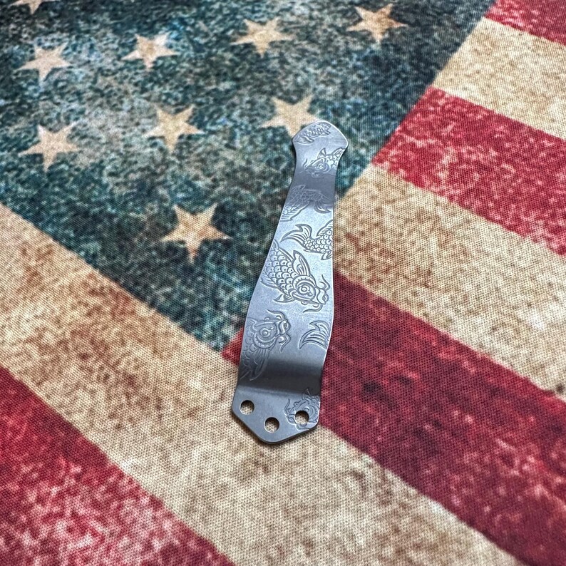 May include: A silver-colored pocket knife clip with an etched koi fish design. The clip has three holes at the base for attachment. The background is an American flag.