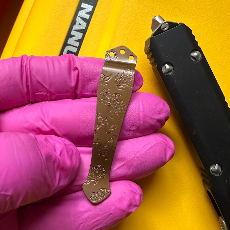 May include: A bronze-colored metal pocket clip featuring an engraved koi fish pattern. The clip has three small circular holes at the top for attachment and a tapered shape. It is held against a bright yellow background next to a black tool.