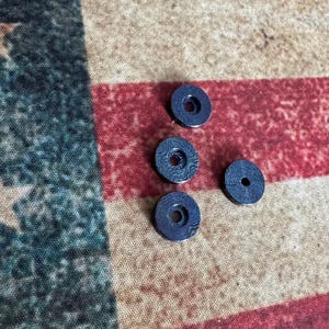Pu&ograve; includere: Quattro piccoli oggetti circolari blu scuro, con un foro centrale e una superficie a motivi, sono disposti su un tessuto a righe rosse, bianche e blu. Gli oggetti sembrano essere componenti decorativi o funzionali.