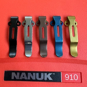 Puede incluir: Cinco clips met&aacute;licos en negro, plateado, bronce, azul y dorado. Cada clip tiene una base curva y una ranura central. La palabra "NANUK" y el n&uacute;mero "910" est&aacute;n impresos en una superficie roja.