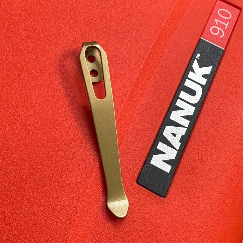 May include: A gold-colored metal clip with two circular holes near the top, next to a black label with the white text "NANUK" and the number "910" in red. The items are on a textured red surface.