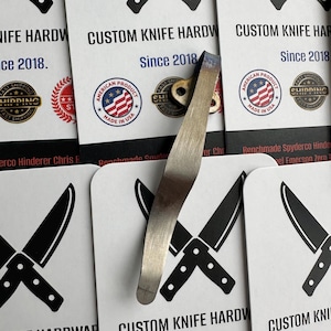 May include: A silver-colored knife hardware component with a curved design, possibly a pocket clip or blade component. The background features cards with the text "CUSTOM KNIFE HARDWARE Since 2018" and an American flag logo.