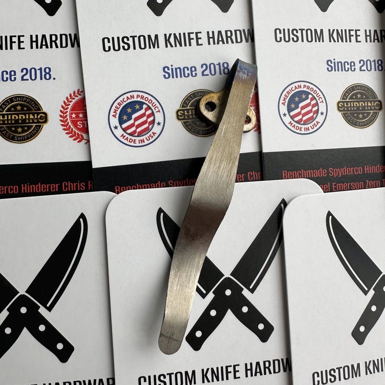 May include: A silver-colored knife hardware component with a curved design, possibly a pocket clip or blade component. The background features cards with the text "CUSTOM KNIFE HARDWARE Since 2018" and an American flag logo.