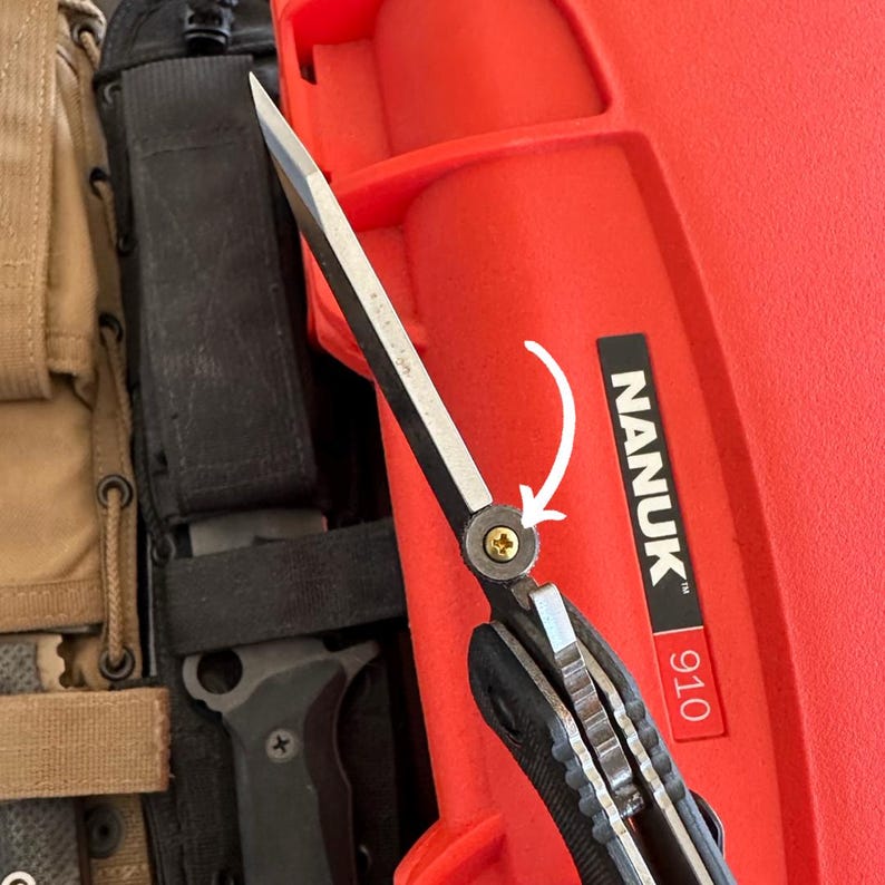 May include: A partially open folding knife with a silver blade and black handle. The knife is positioned in front of a red case with the text "NANUK" and the number "910" printed on it. Other items are visible in the background.