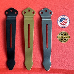 May include: Three metal pocket clips in black, gold, and silver. Each clip has a slotted design with a decorative top featuring three holes and a pointed bottom. The background is red.