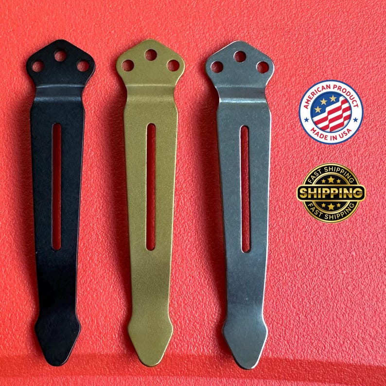 May include: Three metal pocket clips in black, gold, and silver. Each clip has a slotted design with a decorative top featuring three holes and a pointed bottom. The background is red.