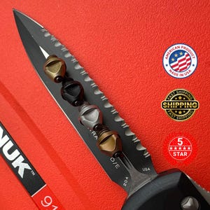 May include: A black and silver knife with a serrated edge and a series of decorative metal beads in gold, black, and silver. The blade has the text "D/E" and "USA". The image also includes a "Made in USA" sticker.