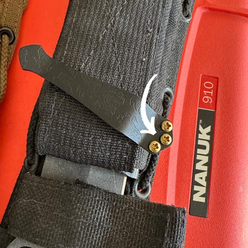 May include: Close-up of a black metal clip with an ornate design, attached to a black fabric sheath. The clip is secured with three gold-colored screws. The sheath is next to a red case with the text "NANUK 910".