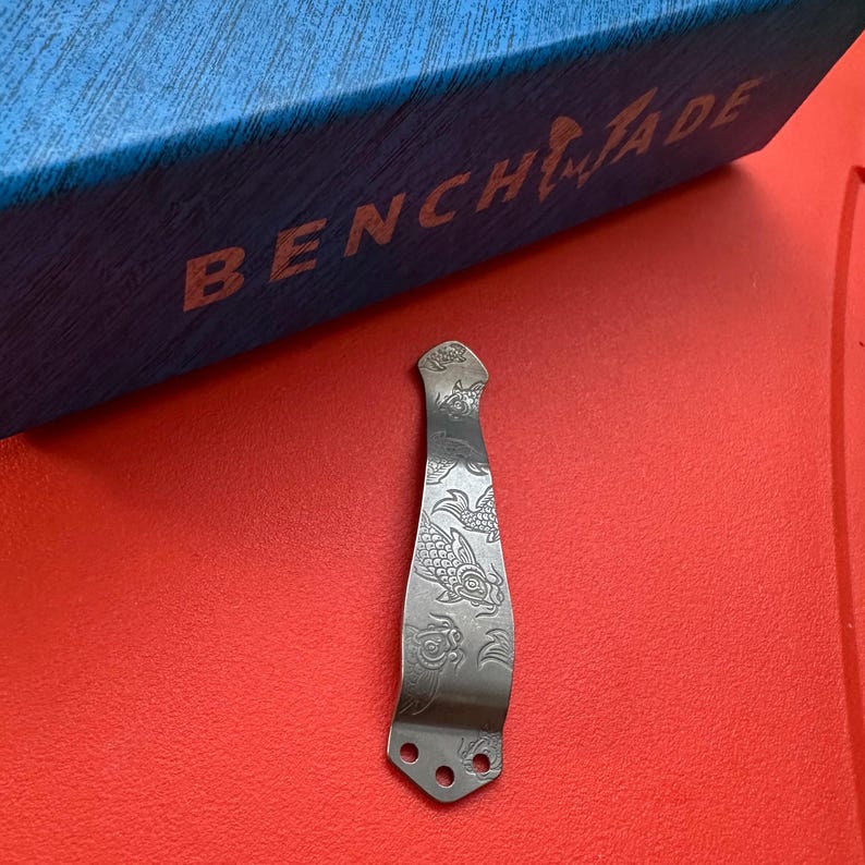 Op de afbeelding: Een zilverkleurige zakmesclip met een gegraveerd koi-visontwerp. De clip heeft drie gaten aan de basis en een gebogen bovenkant. De achtergrond is een felrode ondergrond met een blauwe doos met "Benchmade".