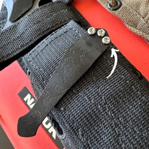 以下が含まれることがあります： 黒い金属製のポケットクリップのクローズアップ。鯉のデザインが施され、3つのシルバーネジで固定されています。 クリップは黒いナイロン製の鞘に取り付けられており、背景は赤色です。