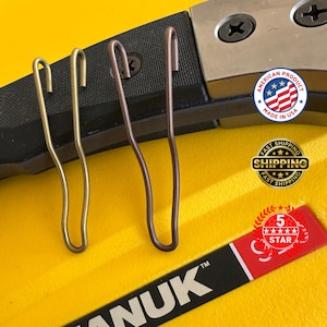 May include: Three metal pocket clips in brass and bronze tones are displayed against a yellow surface. The clips are attached to a black and silver object. The image includes a "Made in USA" sticker and a "5 Star" sticker.