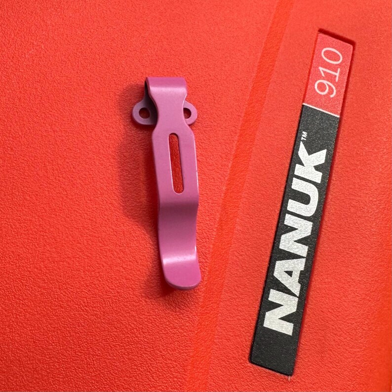 May include: A pink metal clip with a rectangular cutout and two circular holes at the top. The clip is next to a black and red label that reads "NANUK 910". The background is a textured red surface.