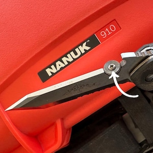 May include: A black and silver Emerson folding knife with a serrated blade, set against a red background. The knife is partially open, showing the blade. The red surface has the text "NANUK" and "910".