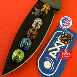 May include: A black knife blade with a series of colorful thumb studs in gold, black, silver, bronze, blue, and green. Includes a tag that says "Genuine AXIS Mechanics" and "Made in USA".