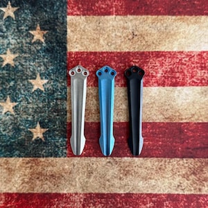 May include: Three metal pocket knife clips in silver, blue, and black, each with a three-hole design at the top. The clips are displayed against a backdrop of the American flag, with its red and white stripes and stars.