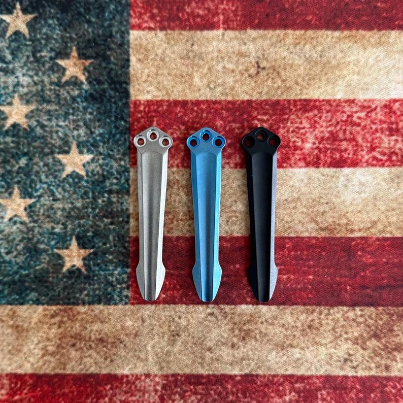 May include: Three metal pocket knife clips in silver, blue, and black, each with a three-hole design at the top. The clips are displayed against a backdrop of the American flag, with its red and white stripes and stars.