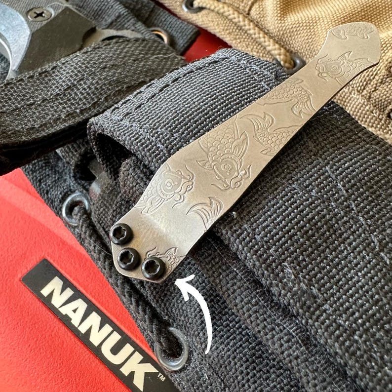 May include: A silver-colored pocket clip with an engraved koi fish design, attached to a black nylon strap. The clip is secured with three black screws. The red case below has the word "NANUK" printed on it.